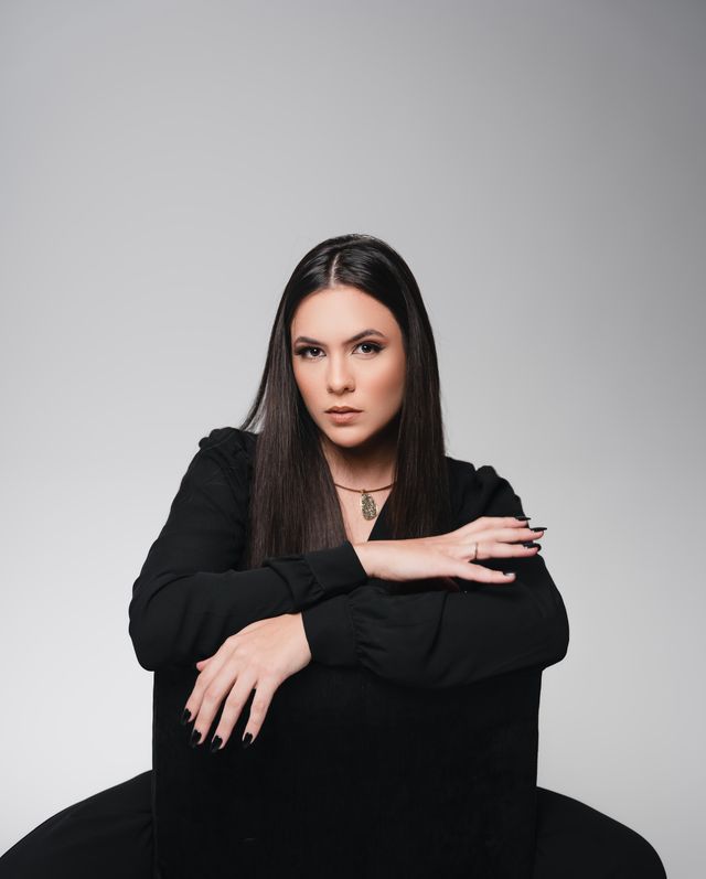 Layza sentada ao contrário numa cadeira, braços cruzados no encosto, olhando para a câmera